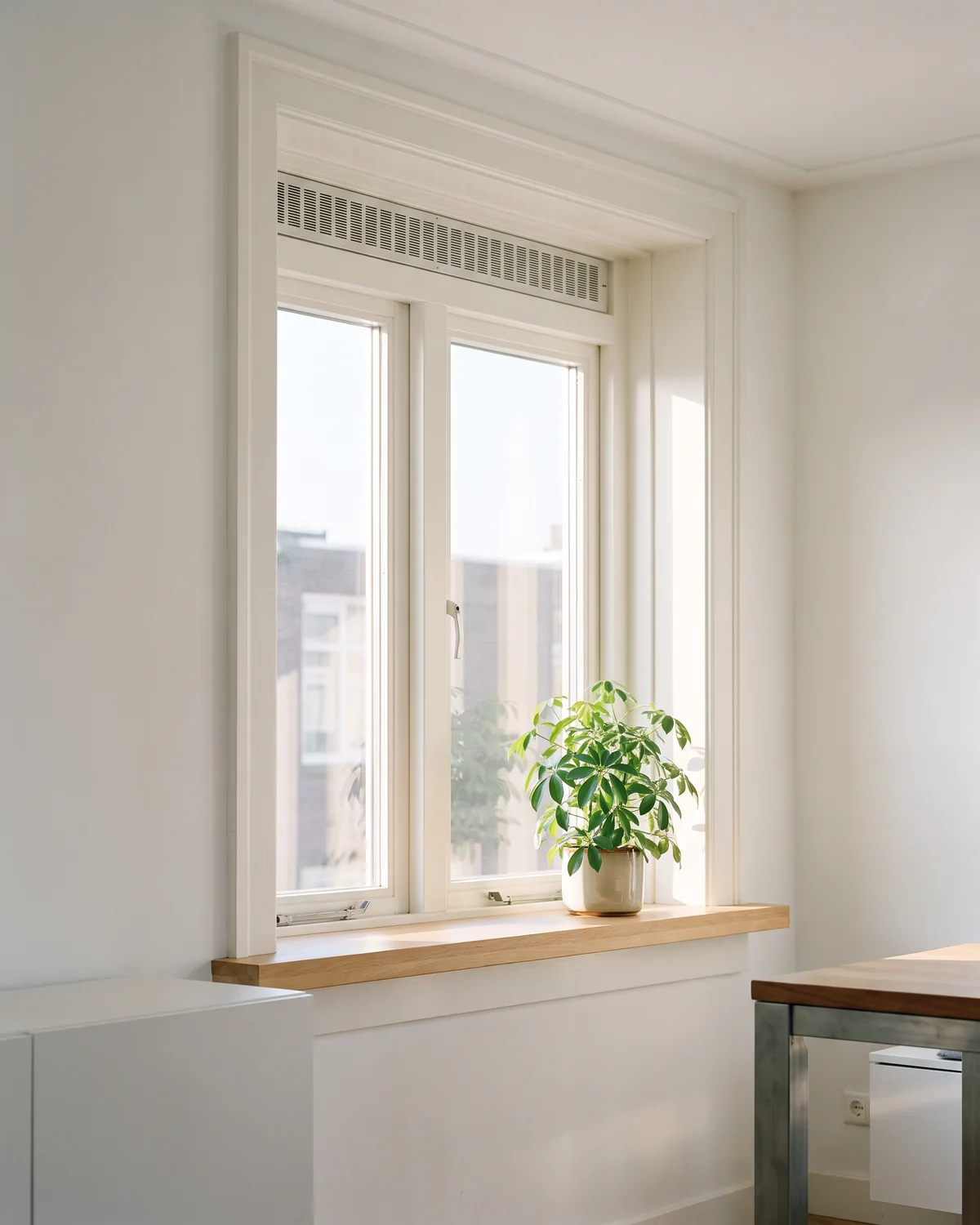 Een helder raam met wit kozijn en een ventilatierooster bovenin, droog glas en zon die naar binnen valt op een vensterbank